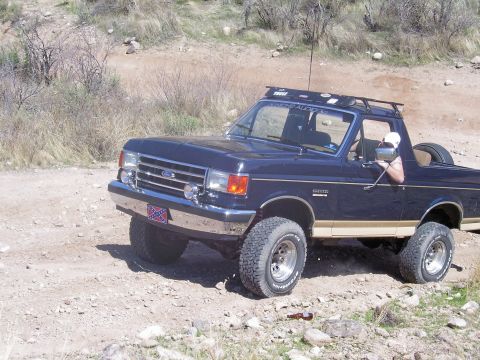 1991 Ford Bronco - A dark blue Ford Bronco with tan trim is seen off-road, equipped with a roof rack and lifted suspension.