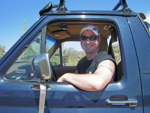 1991 Ford Bronco - A man smiles from the driver's seat of a blue 1991 Ford Bronco parked in a desert landscape.