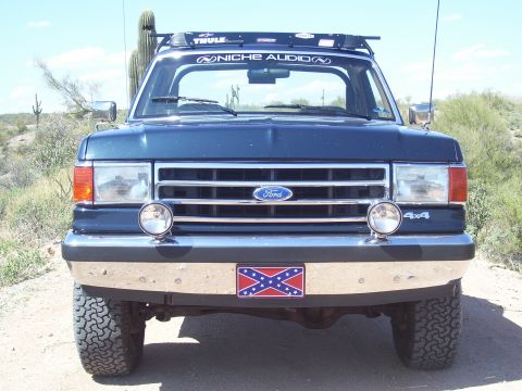 1991 Ford Bronco - Front view of a dark blue Ford Bronco with a chrome bumper and a Confederate flag plate, parked in a desert landscape.