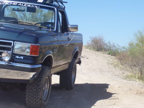1991 Ford Bronco - A 1991 Ford Bronco is seen from a 3/4 front angle on a dusty, off-road path under a clear blue sky.