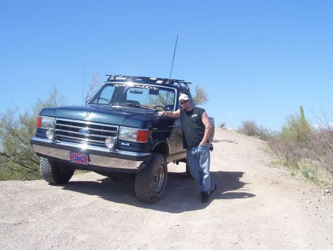 1991 Ford Bronco - A man poses with his lifted Ford truck on a dirt road under a clear blue sky.