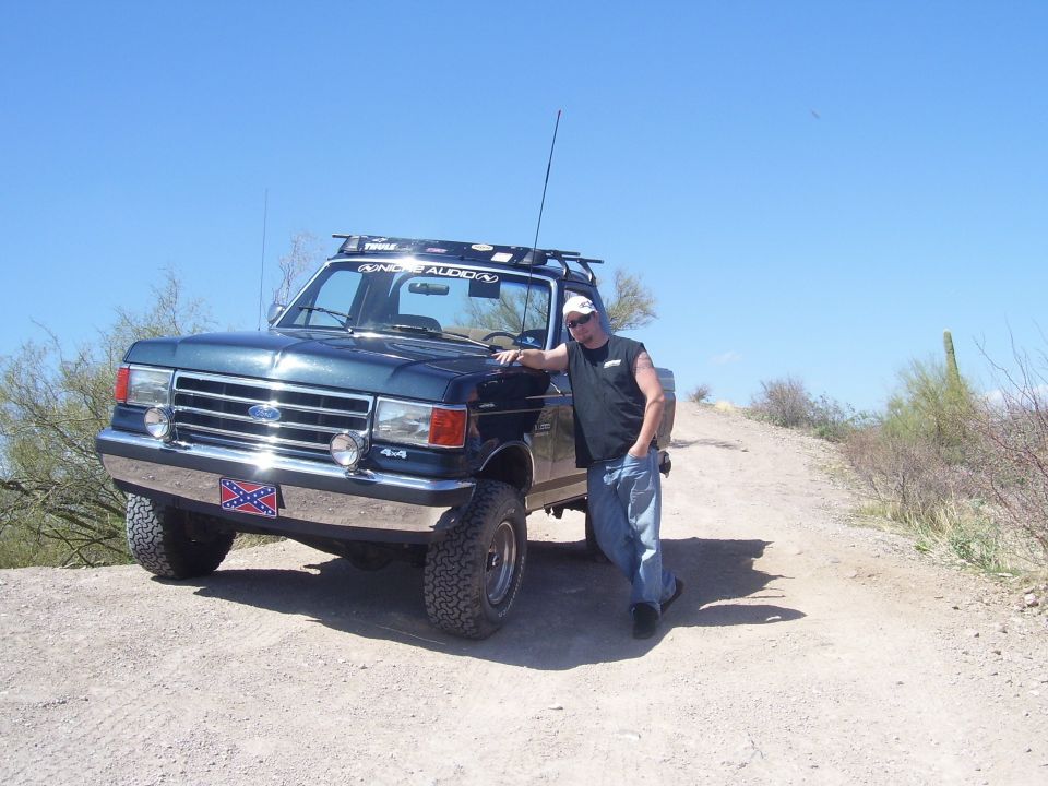Guy leaning on dark green Ford F-Series truck with roof rack and a rebel flag plate on the bumper