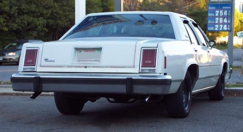 1985 Ford Crown Victoria LTD - A white sedan is parked near a gas station with visible gas prices.