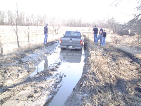 1981 Ford Bronco - Rear view of a gray Ford pickup sitting in a water-filled mud trench cut through a winter field, with three bystanders standing on the bank…