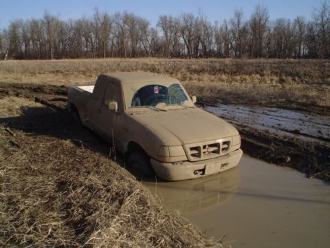 1981 Ford Bronco - A pickup truck is stuck in a muddy ditch, with its tires submerged in water and mud.