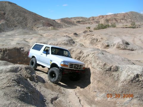 1985 Ford Bronco - A white Ford Bronco tackles a challenging off-road trail in a dry, rocky environment.
