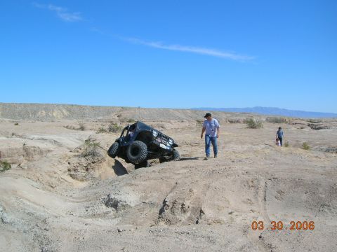 1985 Ford Bronco - A black off-road vehicle attempts to navigate a steep dirt obstacle in a desert environment under a clear blue sky.