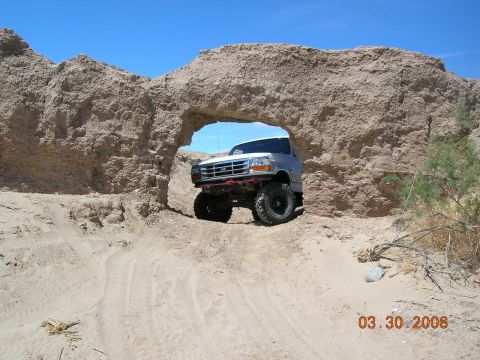 1985 Ford Bronco - A lifted Ford Bronco navigates a natural rock arch in a sandy, arid environment.