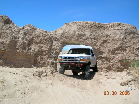 1985 Ford Bronco - White Ford Bronco navigates a desert rock archway on a sunny day.