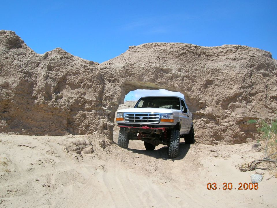 White Ford Bronco driving through a natural rock archway in a desert landscape