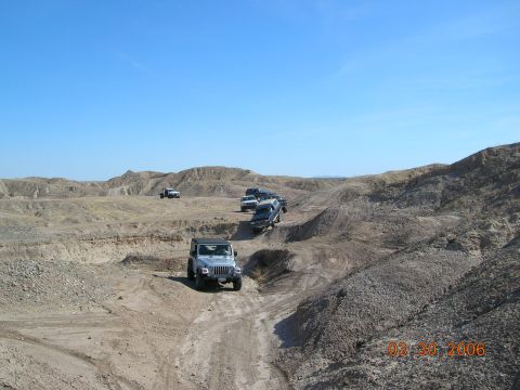 1985 Ford Bronco - Multiple Jeeps traverse a desert landscape on a sunny day, with tire tracks visible on the dirt path.