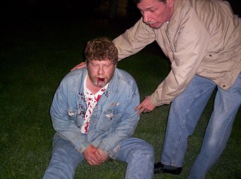 1989 Ford Bronco - A man with a bloody face and shirt is attended to by another man on a grassy area at night.