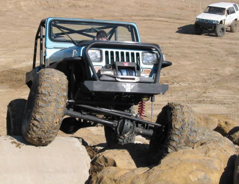 1981 Ford Bronco - A modified blue Jeep Wrangler navigates a rocky obstacle course, with another SUV visible in the distance.