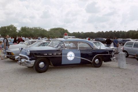 1991 Ford Crown Victoria - This vintage police car, likely a 1956 model, is parked outdoors with other vehicles and onlookers in the background.
