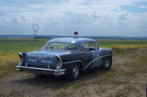 1991 Ford Crown Victoria - A 1956 Buick Century police car is parked on a dirt road in a grassy field under a cloudy sky.