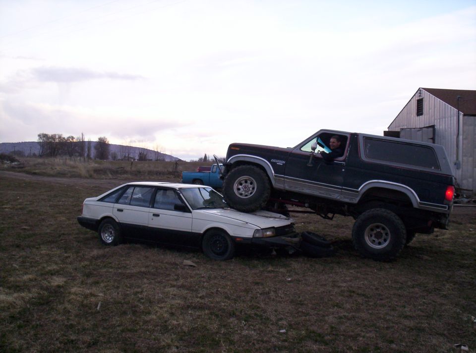 Lifted black Ford Bronco parked with its front wheels up on the hood of a wrecked white sedan in a field
