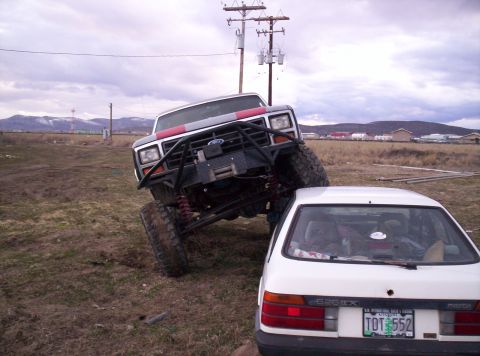1984 Ford Bronco - A heavily modified Ford Bronco is positioned precariously over a white Mazda sedan in a rural setting.