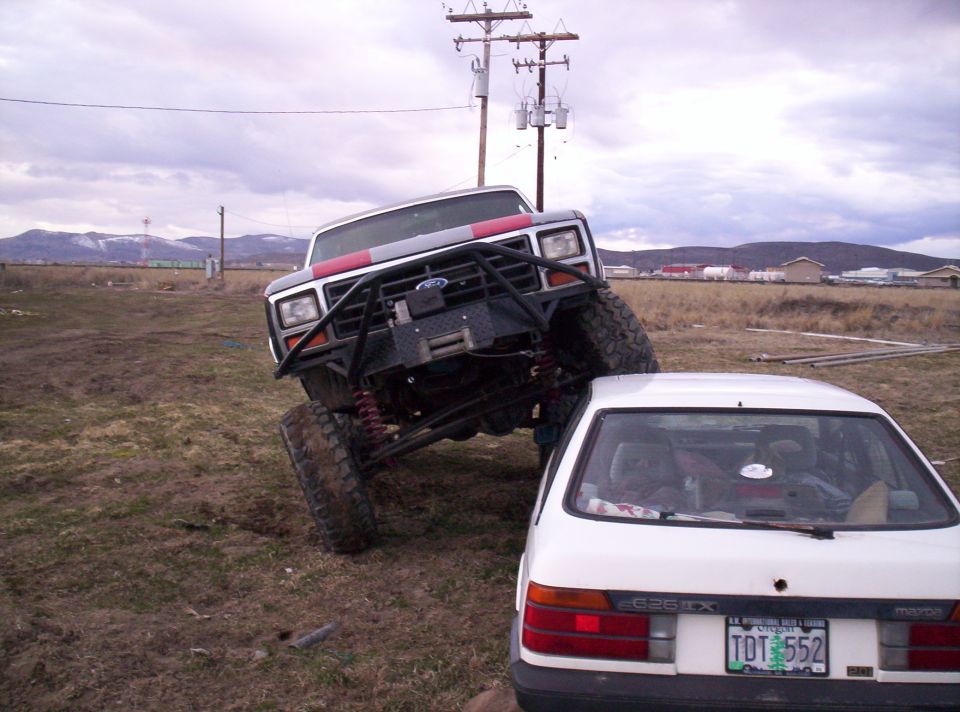 Modified Ford Bronco, dark gray with red stripes, on a custom bumper, angled over a white Mazda sedan