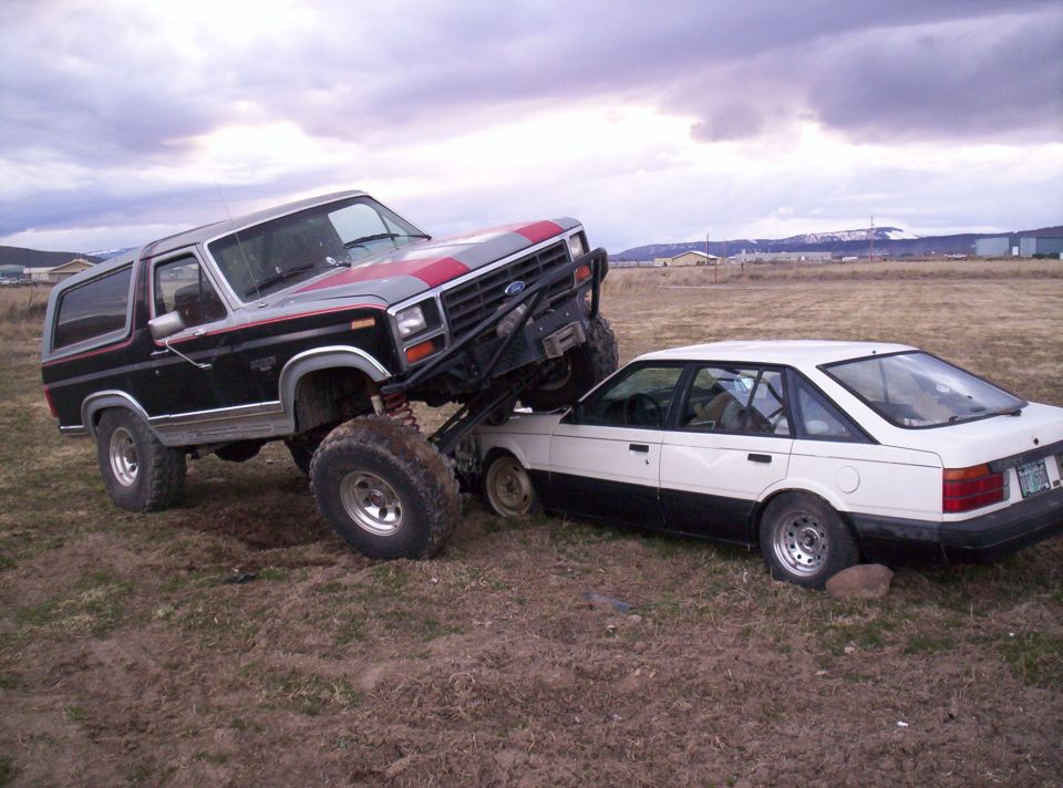 Black and red lifted Ford Bronco with front wheels driven up onto a white sedan in a grassy field