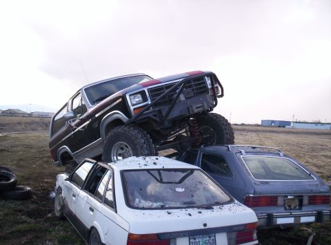 1984 Ford Bronco - A heavily modified SUV is piled on top of two other vehicles in a junkyard setting.
