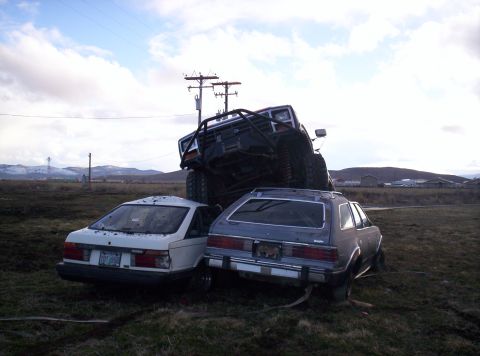 1984 Ford Bronco - An off-road vehicle is precariously balanced on top of two other vehicles in a grassy field under a cloudy sky.
