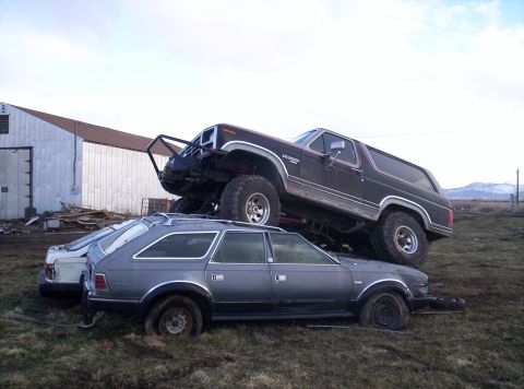 1984 Ford Bronco - A lifted Ford Bronco is stacked on top of two older station wagons in a junkyard setting.