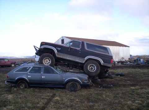 1984 Ford Bronco - A lifted Ford Bronco is parked on top of a station wagon in a junkyard setting.
