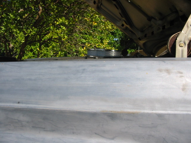 Engine bay with hood open, showing air filter and some engine components. Gray metal surface in foreground