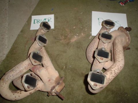 1981 Ford Bronco - A pair of used V8 cast-iron exhaust manifolds sitting on green carpet, coated in off-white high-heat paint with rust bleeding through and h…