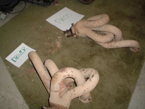 1981 Ford Bronco - Two exhaust manifolds, labeled 'DRIVER' and 'PASS', lie on a carpeted floor.
