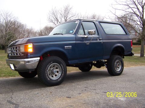 1987 Ford Bronco - A blue Ford Bronco is shown from the side and front, parked on a road with trees in the background.