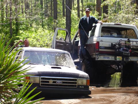 1989 Ford Bronco - Two Ford Broncos are stuck in the mud in a wooded area, with people standing on them.