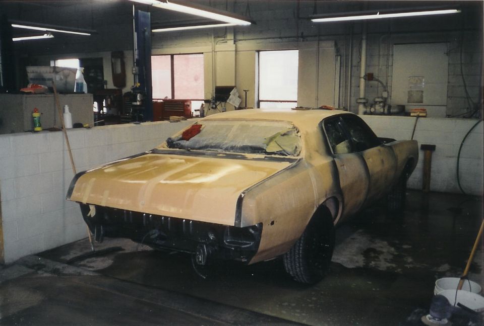 Tan primer two-door coupe in body shop, rear 3/4 view, taped windows, mid-restoration