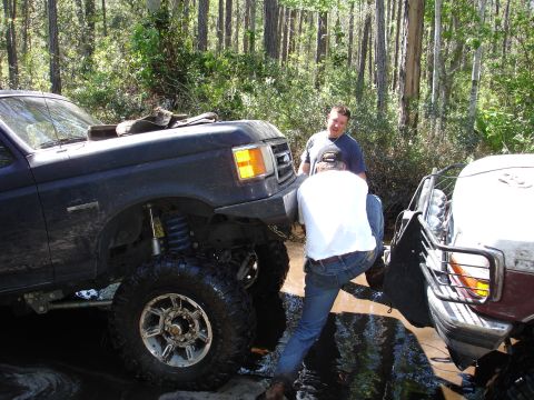 1989 Ford Bronco - Two men attempt to free a dark blue lifted truck stuck in muddy water, with another vehicle partially visible to the right.