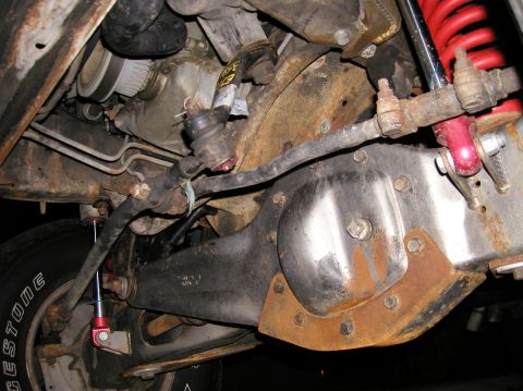 1984 Ford Bronco - Underside view of a solid front axle showing the differential housing with a bolt-on cover and rusty skid/truss plate, tie rod and drag lin…