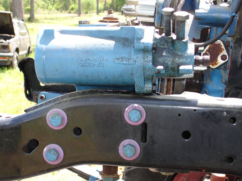 Blue hydraulic pump assembly with part number E10-1040-JB, mounted on a black frame with pink and blue bolts. Another vehicle is visible in the background