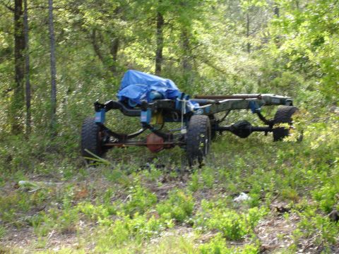 1989 Ford Bronco - The chassis of a vehicle, including its frame and axles, is visible in a wooded area.