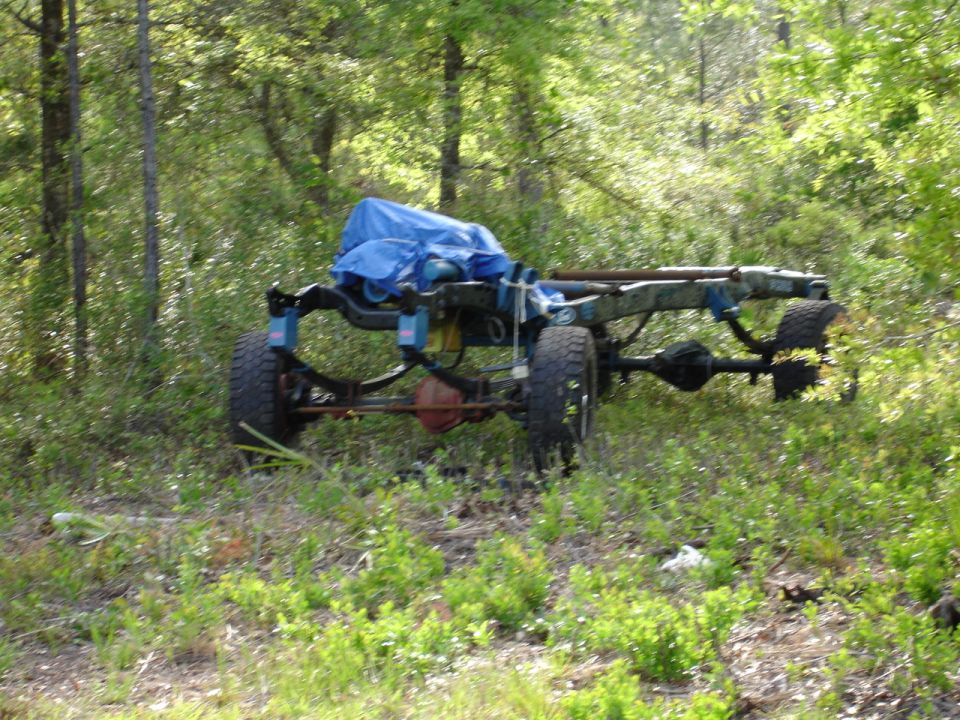 vehicle frame with axles and wheels, covered with a blue tarp, sitting in grassy woods