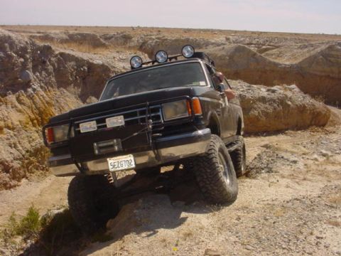 1988 Ford Bronco - This black Ford Bronco is shown off-roading on a dirt path with large tires and auxiliary lights.
