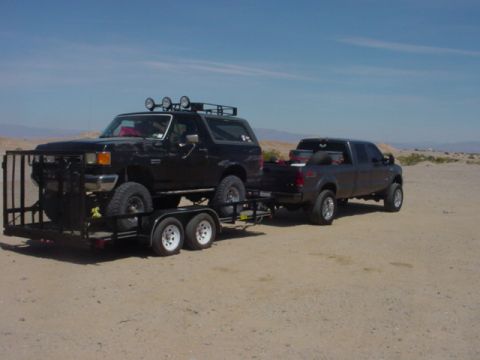 1988 Ford Bronco - A black Ford Bronco is loaded onto a trailer and towed by a pickup truck across a dry, dusty terrain under a clear blue sky.