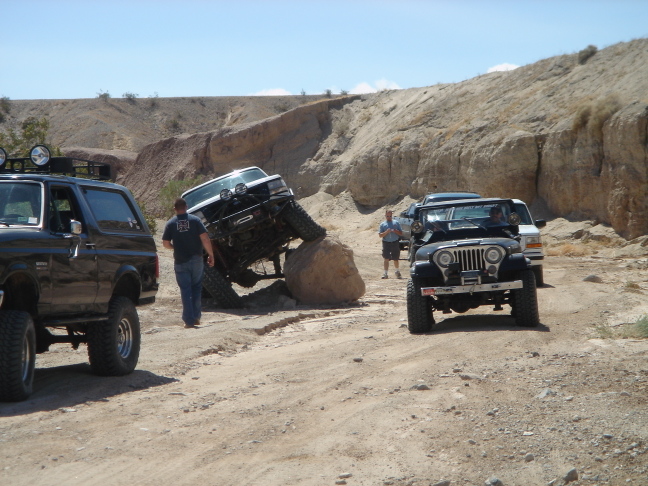 Black 1988 Ford Bronco parked on a dirt trail next to a white Bronco climbing a rock