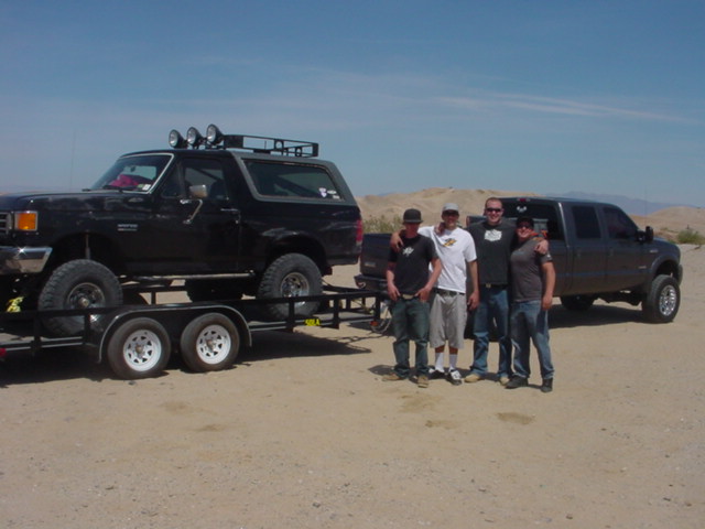 Black Ford Bronco on a trailer, 3/4 front view. It has a roof rack with lights. A dark gray Ford F-250 Super Duty is parked behind it