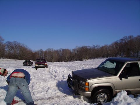 2000 Chevrolet Tahoe - A silver SUV is stuck in deep snow, with two individuals attempting to assist. Another vehicle is visible in the background.