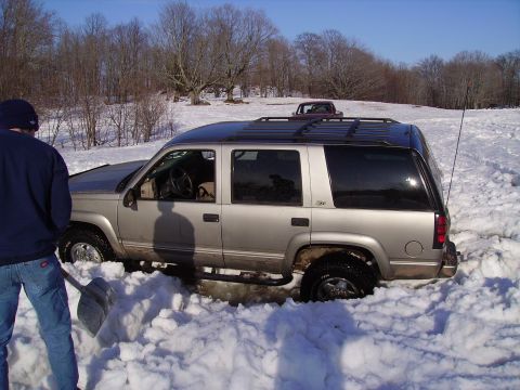 2000 Chevrolet Tahoe - A person attempts to dig out a silver SUV that is stuck in deep snow, with another vehicle visible in the distance.