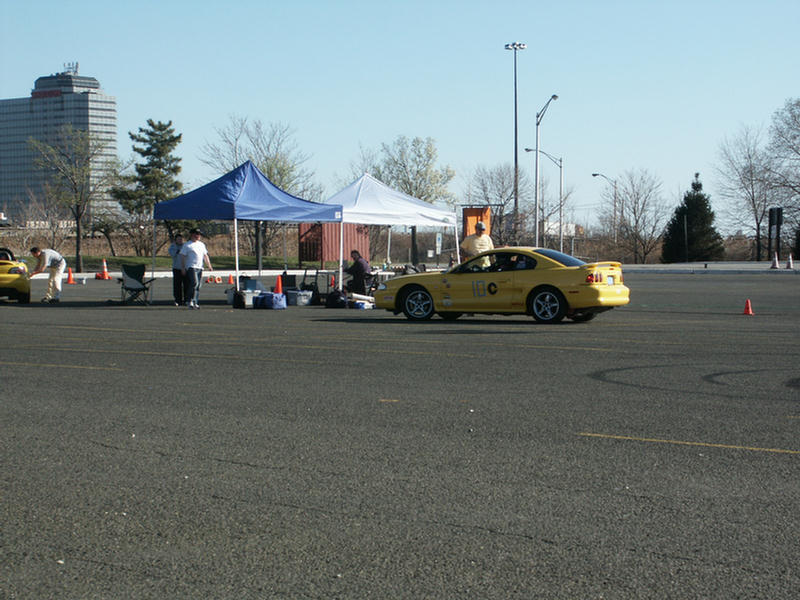 Yellow coupe with racing number 10 on door, parked on asphalt with tents and people nearby