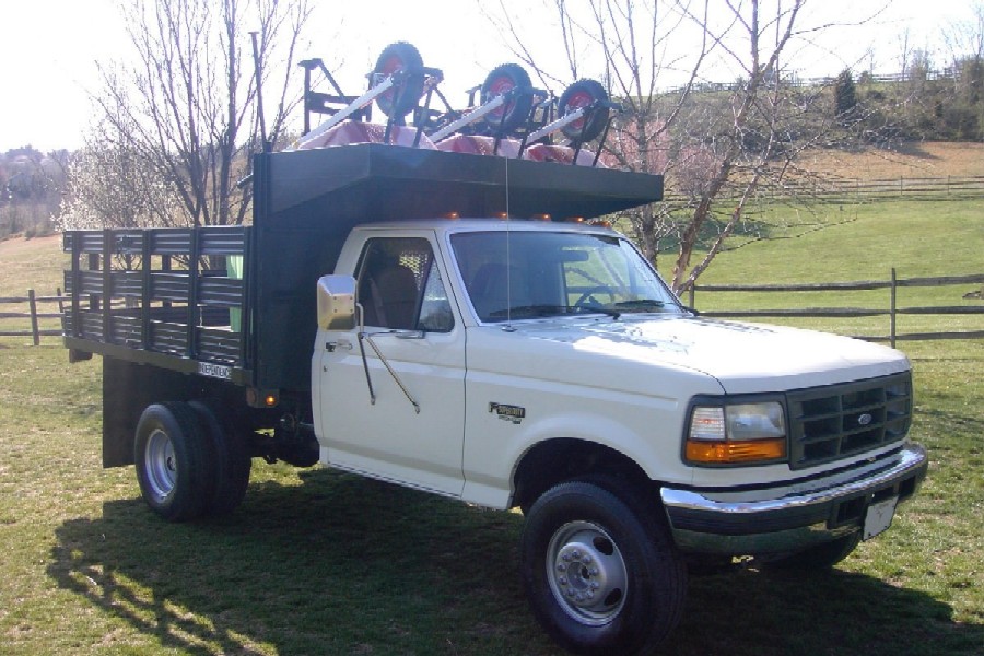 White Ford dually flatbed truck with stake sides and equipment on an overhead rack in a green field