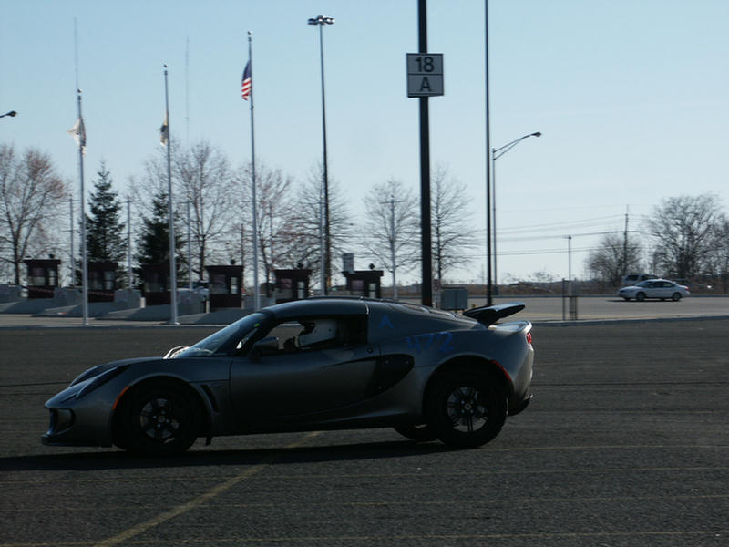 Silver Lotus Elise driving on pavement, flags and light poles in background