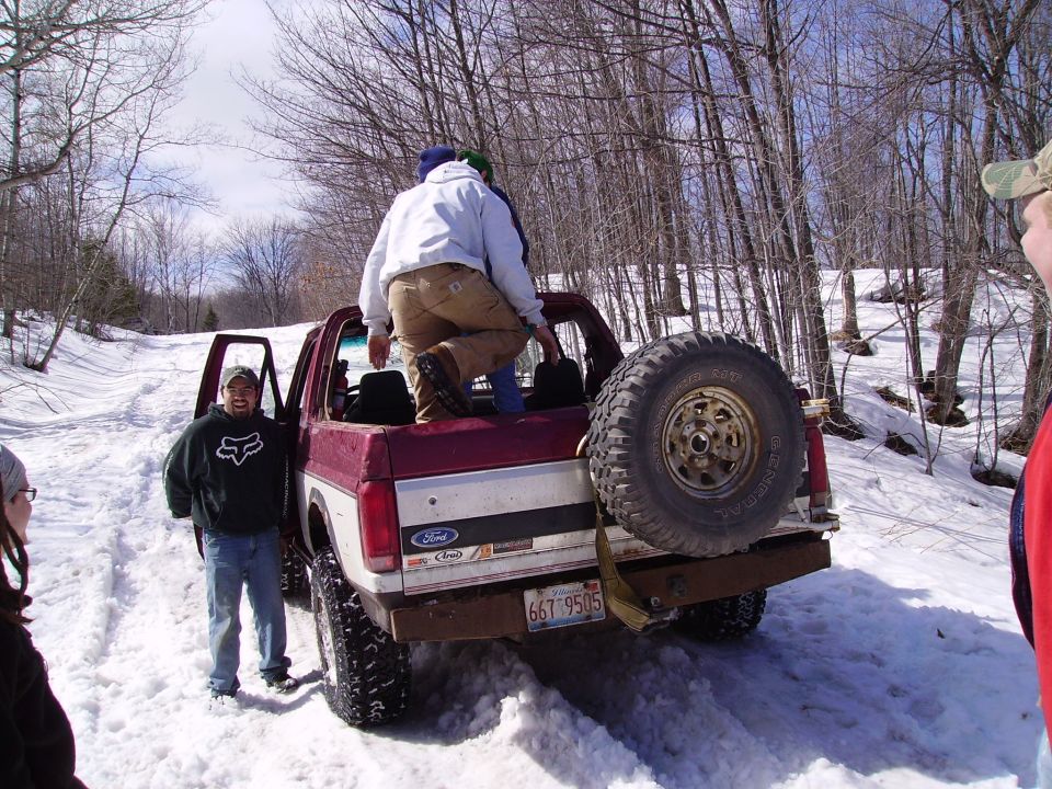 Ford pickup truck with spare tire mounted on rear, people in bed, in snowy woods. License plate reads 667 9505