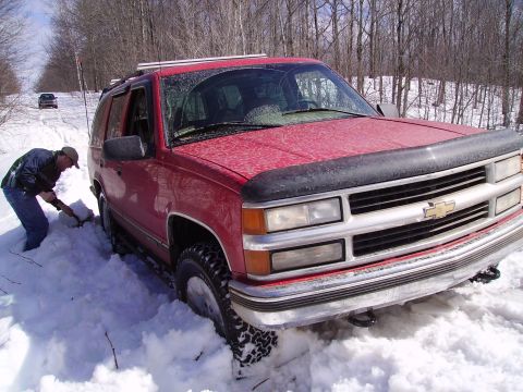 2000 Chevrolet Tahoe - A red Chevrolet Tahoe is stuck in deep snow while a person attempts to dig it out, with another vehicle in the distance.