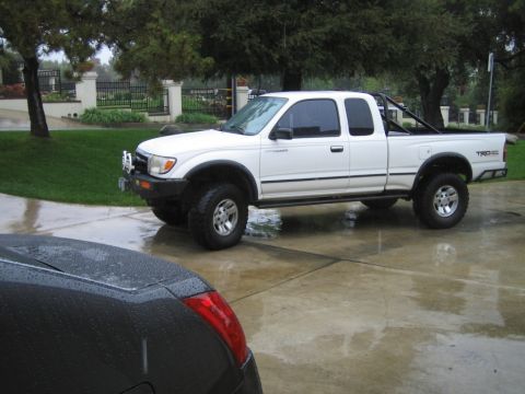 1985 Ford Bronco - White Toyota Tacoma pickup truck with TRD badging parked on a wet driveway, with a black car in the foreground.